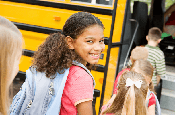 Young girl wearing a backpack smiles while stepping onto a yellow school bus.