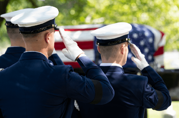 Three uniformed military personnel salute a flag-draped coffin during a solemn outdoor funeral.