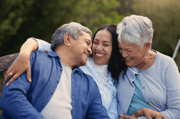 Adult daughter sits between her parents on a bench outdoors, smiling and embracing them as they laugh together, symbolizing a loved one who may one day serve as a life insurance beneficiary.