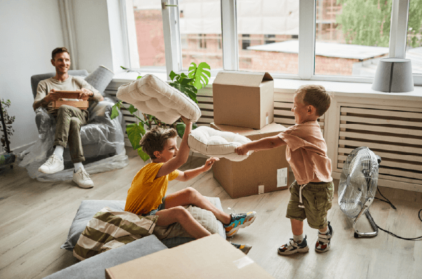 Children play with pillows in a room filled with moving boxes while an adult begins unpacking, capturing the excitement and transition of moving into a new home during a military PCS.