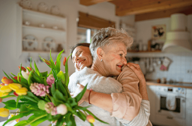 Two people embrace in a cozy kitchen while one holds a bouquet of fresh flowers, reflecting the loved ones who may one day become a life insurance beneficiary.