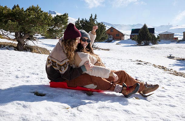 Bundled in winter gear, two women sled down a hill with smiles and laughter, showcasing the fun and connection that snowy getaways can bring.